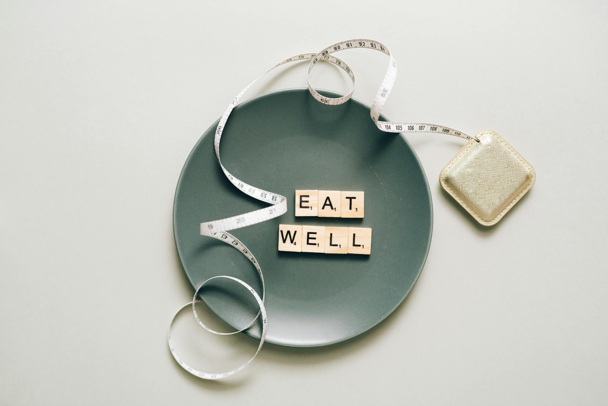Flat lay of scrabble tiles spelling 'eat well' on a plate with a tape measure, promoting healthy eating.